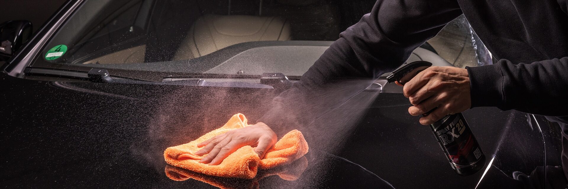 Een cardetailer is met een oranje microvezeldoek de buitenkant van de auto aan het reinigen, met behulp van RIWAX Spray Finish RX 20.